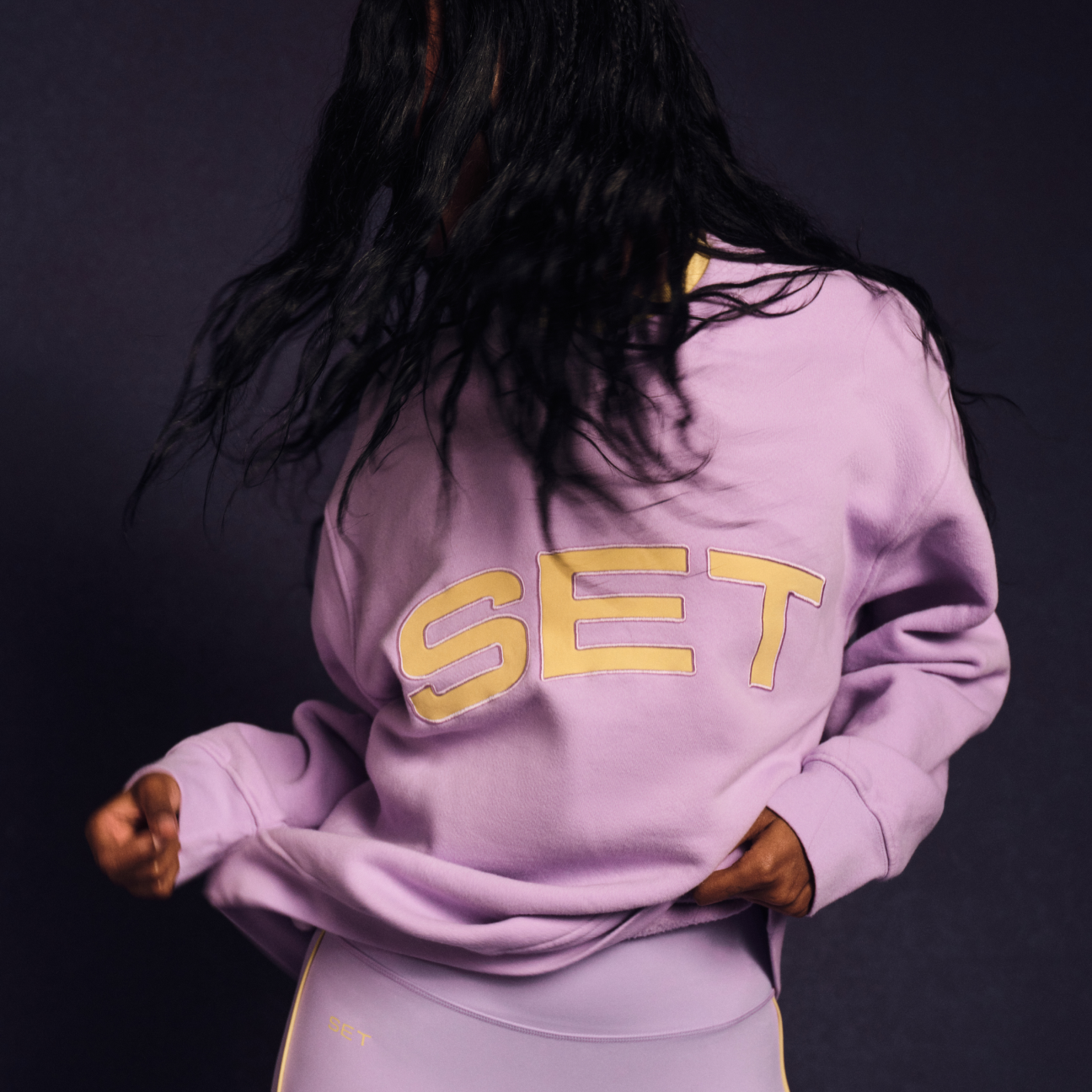 Person wearing a purple hoodie with 'SET' printed on the back against a dark background