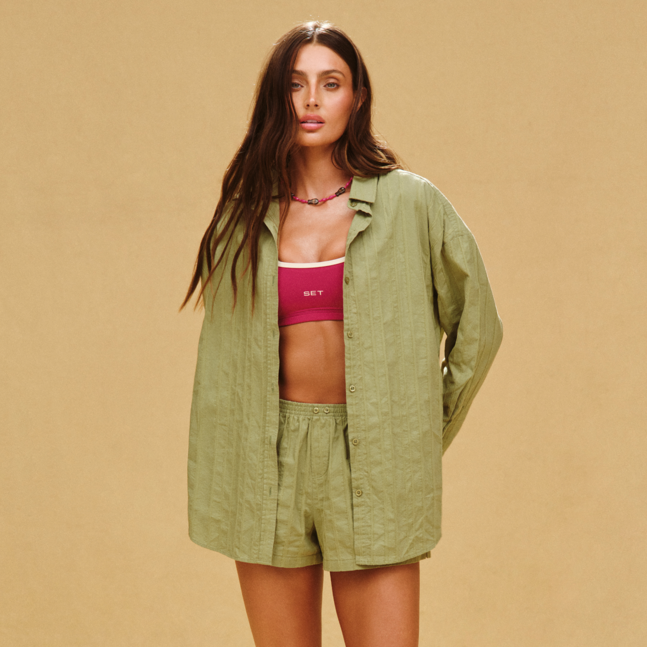 Woman wearing a green shirt and shorts over a pink sports bra on a beige background