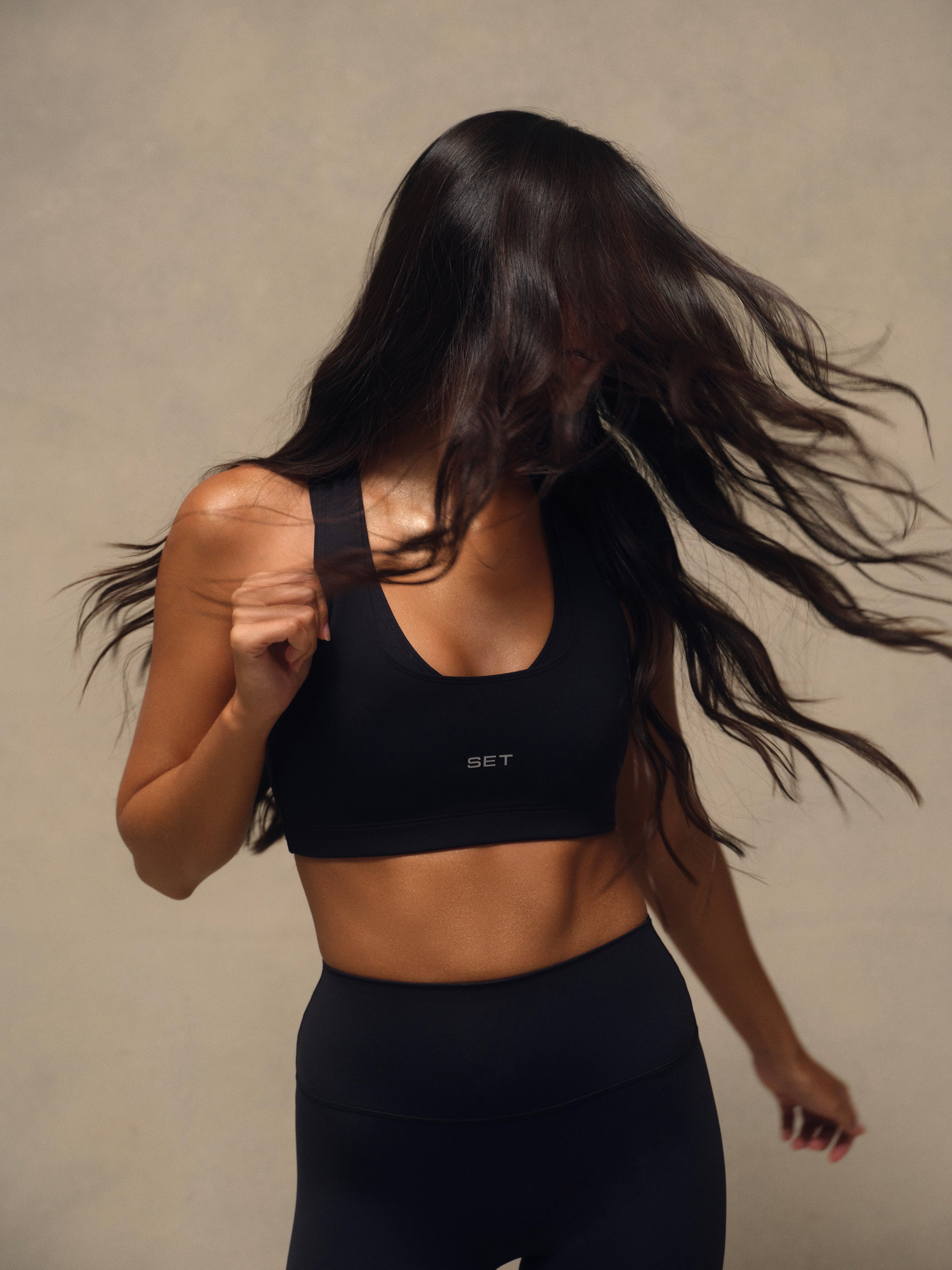 A woman wearing a black sports bra and matching black leggings, captured mid-motion with her long dark hair blowing across the frame against a plain beige studio background. The sports bra has a small visible 'SET' logo on the front.