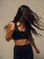 A woman wearing a black sports bra and matching black leggings, captured mid-motion with her long dark hair blowing across the frame against a plain beige studio background. The sports bra has a small visible 'SET' logo on the front.