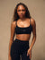 A woman modeling a black athletic sports bra and matching high-waisted leggings against a neutral beige studio backdrop. The sports bra has thin straps and a small white 'SET' logo on the center chest. 