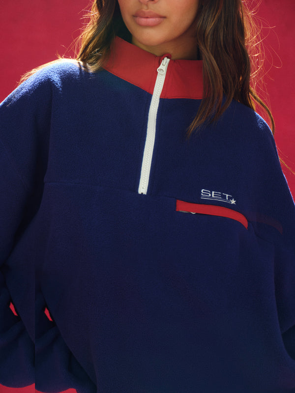 Navy blue pullover with red collar and white zipper, featuring a logo on the chest against a red background.