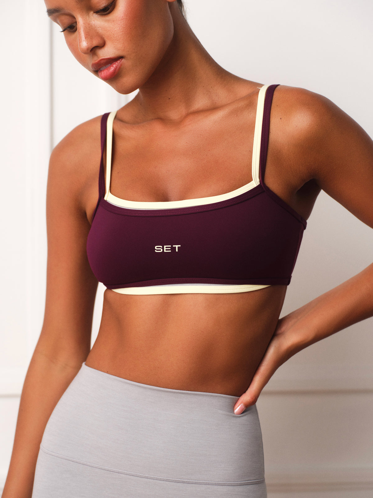 A woman wearing a deep burgundy sports bra with cream trim and the brand name 'SET' printed on the front, paired with light gray leggings. 