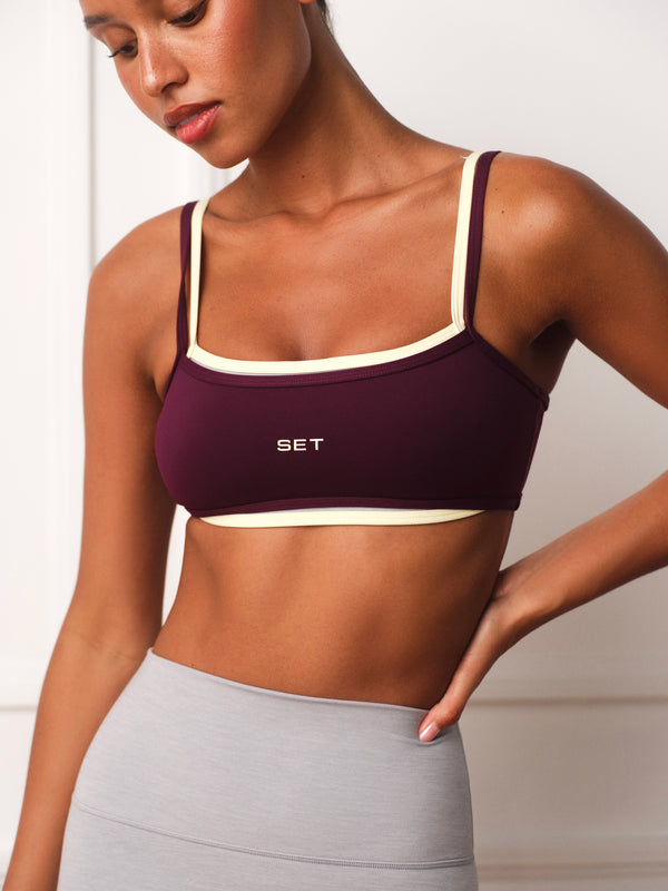 A woman wearing a deep burgundy sports bra with cream trim and the brand name 'SET' printed on the front, paired with light gray leggings.