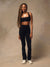 A woman modeling a matching black activewear set consisting of a square-neck sports bra and high-waisted leggings, paired with white sneakers, against a textured beige studio backdrop.