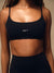 A close-up of a model wearing a black athletic sports bra with thin straps and a scoop neckline, featuring the word 'SET' printed in white on the center chest. The matching black athletic bottoms are partially visible at the waist. <model-info>Lena wears a size XS and is 5’7”, 24½” waist, 31" bust.</model-info>