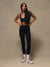 A woman modeling a black sports bra and matching black high-waisted leggings, paired with white socks and white sneakers, standing against a textured beige studio backdrop. <model-info>Jelene wears a size XS and is 5’7”, 23” waist, 32AB bust.</model-info>