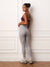 A woman stands against a white paneled wall wearing light gray high-waisted leggings, a dark burgundy sports bra, white socks, and gray sneakers.