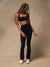 A woman with long blonde dreadlocks is shown from behind wearing a matching black activewear set consisting of a sports bra and high-waisted leggings, paired with white sneakers, against a textured beige studio backdrop.