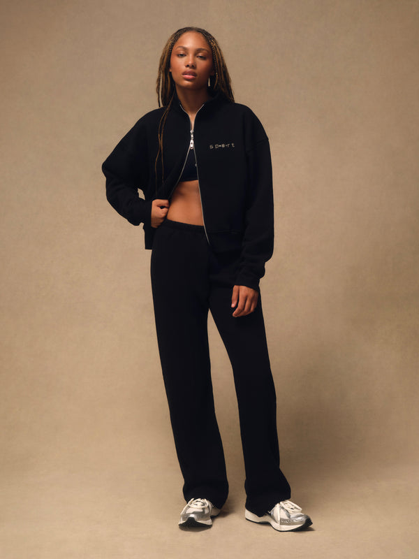 A model stands against a warm beige studio backdrop wearing a black zip-up jacket with a small white embroidered logo on the chest, black cropped tank top, black wide-leg sweatpants, and white sneakers. 