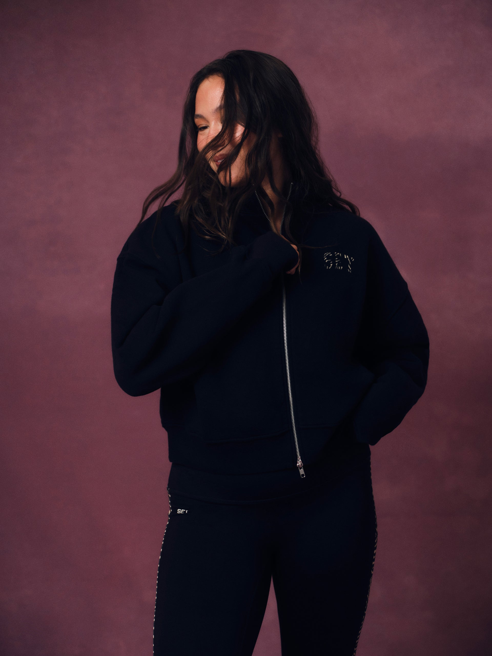 A woman wearing a dark navy zip-up hoodie with 'SET' embroidered on the chest and matching dark pants, posing against a textured mauve-pink studio backdrop. <model-info> Christina wears a size L and is 5’9, 29½” waist, 36" bust.</model-info> 