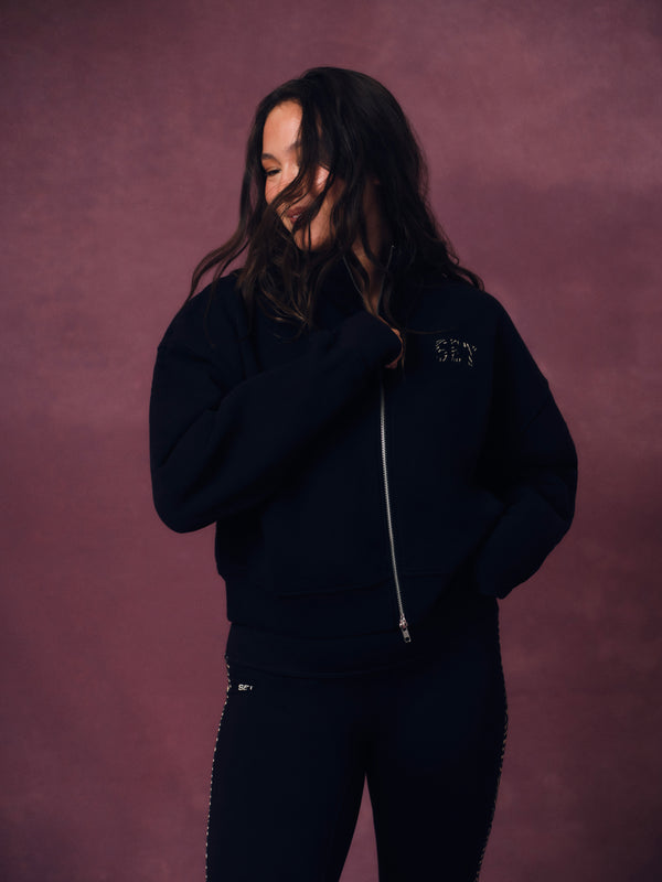 A woman wearing a dark navy zip-up hoodie with 'SET' embroidered on the chest and matching dark pants, posing against a textured mauve-pink studio backdrop. 