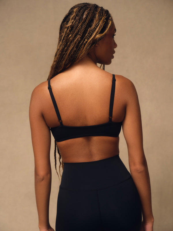 A woman shown from the back wearing a black sports bra and matching black high-waisted leggings against a warm beige studio background.