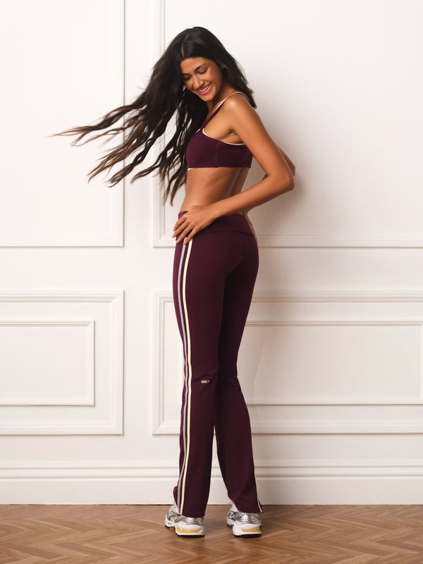 A woman modeling a matching burgundy activewear set consisting of a sports bra and high-waisted track pants with white side stripes, standing against a white paneled wall.