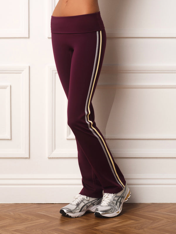 A person wearing burgundy athletic leggings with a double stripe down the side, paired with white and gray sneakers, standing against a white paneled wall.