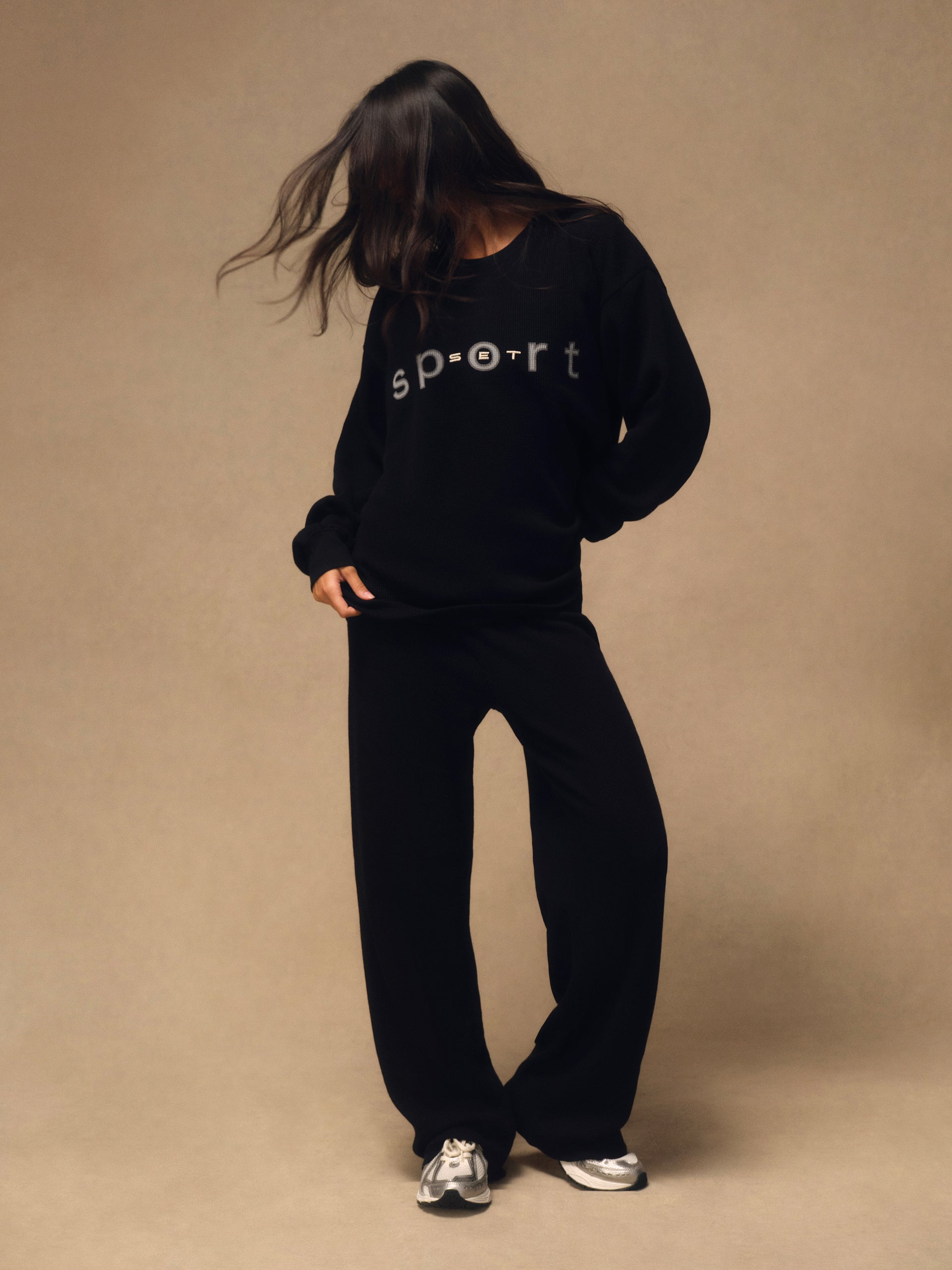 A model wearing a black sweatshirt and matching sweatpants set, featuring the word 'sport' printed in white across the chest, posed against a solid beige studio backdrop.