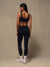 A woman is shown from behind wearing a matching black activewear set consisting of a sports bra and high-waisted leggings, paired with white socks and sneakers, against a textured beige studio backdrop.