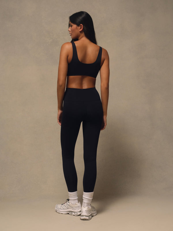 A woman is shown from behind wearing a matching black activewear set consisting of a sports bra and high-waisted leggings, paired with white socks and sneakers, against a textured beige studio backdrop.