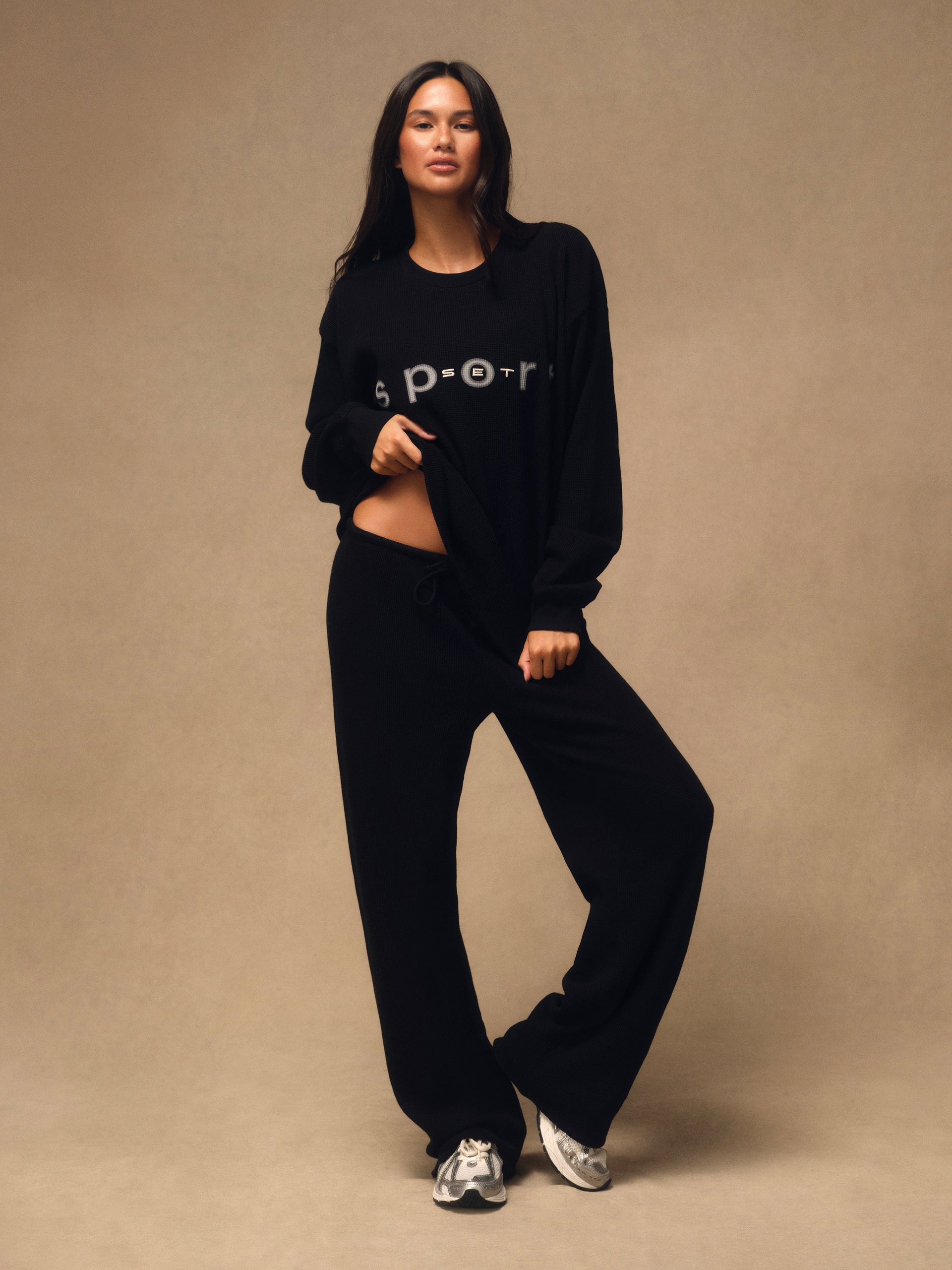 A woman modeling a black cropped long-sleeve sweatshirt with 'sport' printed in white across the chest, paired with matching black wide-leg sweatpants and silver sneakers, against a solid beige studio backdrop. <model-info>Jelene wears a size XS and is 5’7”, 23” waist, 32AB bust.</model-info>