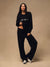 A woman modeling a black cropped long-sleeve sweatshirt with 'sport' printed in white across the chest, paired with matching black wide-leg sweatpants and silver sneakers, against a solid beige studio backdrop. <model-info>Jelene wears a size XS and is 5’7”, 23” waist, 32AB bust.</model-info>
