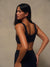 A woman wearing a black sports bra and matching black leggings, photographed from the back against a neutral beige studio backdrop.