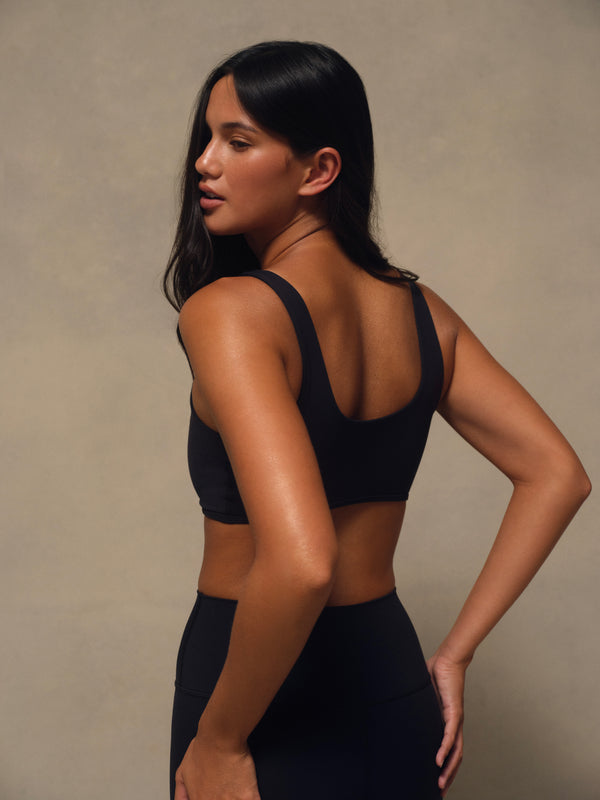 A woman wearing a black sports bra and matching black leggings, photographed from the back against a neutral beige studio backdrop.