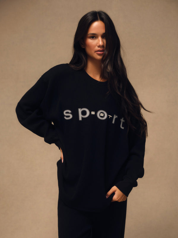 A woman modeling a black crewneck sweatshirt with 'sport' printed in white across the chest, posed against a neutral beige background. 
