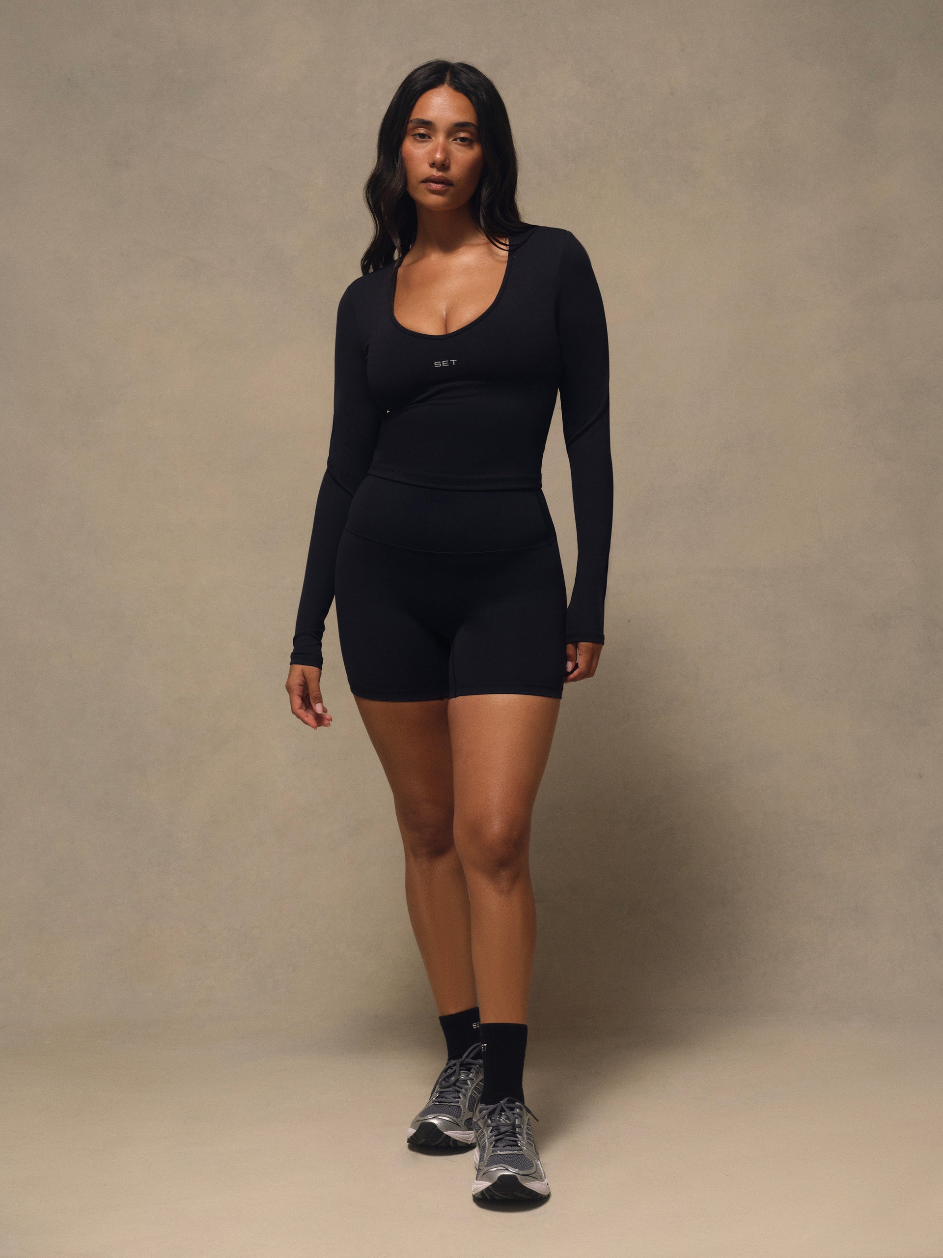 A woman modeling a black long-sleeve athletic long sleeve with matching black shorts, styled with black socks and gray running shoes against a neutral beige studio backdrop.