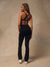 A woman stands with her back to the camera wearing a black strappy athletic top and black high-waisted pants against a textured beige studio backdrop.