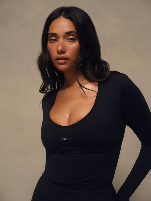 A woman with long dark hair wearing a tight-fitting black long-sleeve scoop-neck top with a small white 'SET' logo on the chest, posed against a neutral textured beige background.