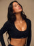 A woman wearing a black long-sleeve crop top with a twisted front detail and a scoop neckline, featuring the brand name 'SET' printed in white on the lower right side. She is posing against a neutral beige background.