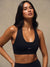 A woman wearing a black athletic sports bra with a small silver 'SET' logo on the front, posed against a neutral beige studio background. <model-info>Jelene wears a size XS and is 5’7”, 23” waist, 32AB bust.</model-info>