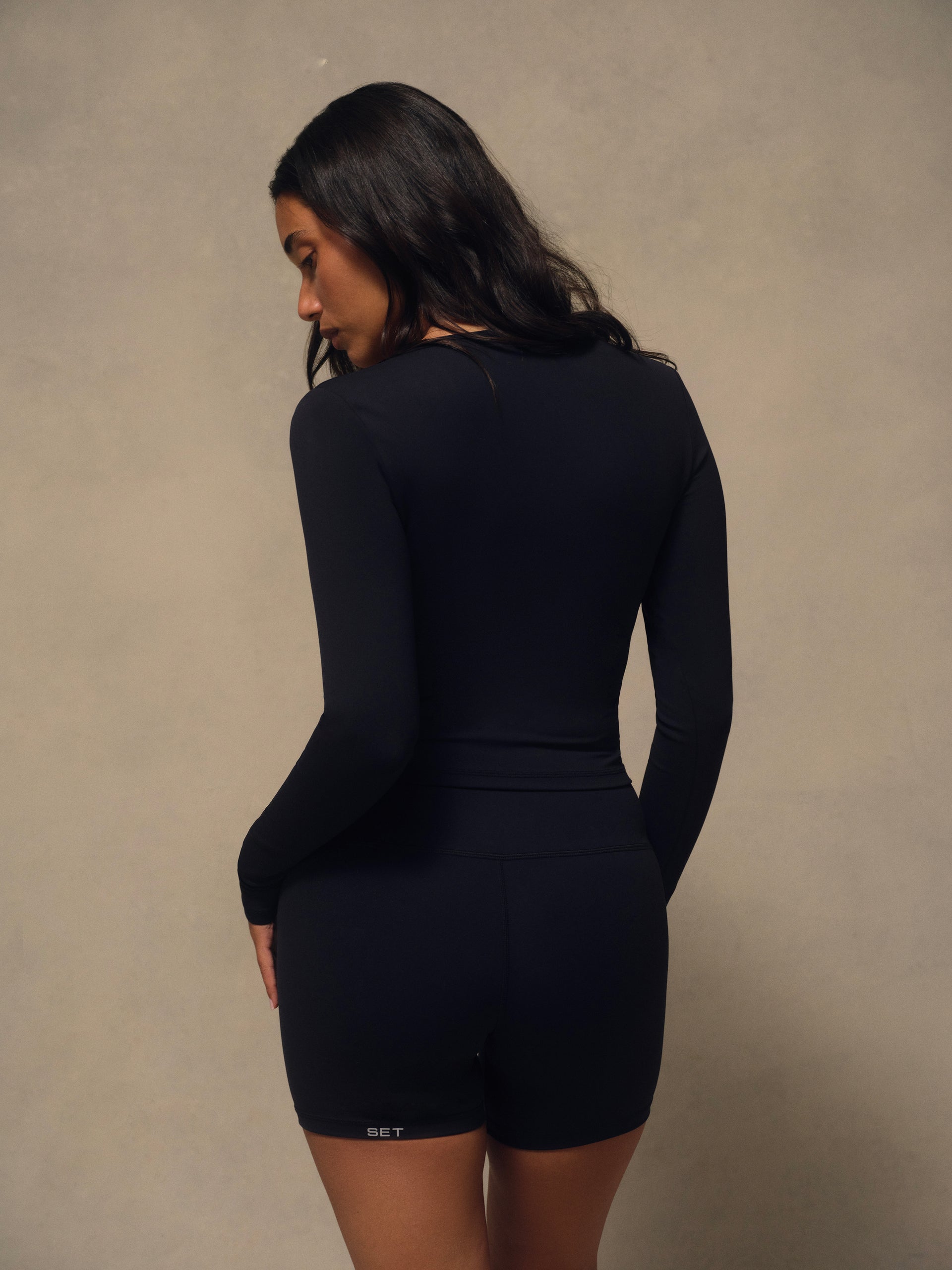 A woman wearing a black long-sleeve athletic top, shown from the back against a neutral textured beige background.