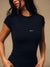 A woman wearing a fitted black short-sleeve athletic top with a small white 'SET' logo on the chest, photographed against a warm beige studio background. <model-info>Jelene wears a size XS and is 5’7”, 23” waist, 32AB bust.</model-info>