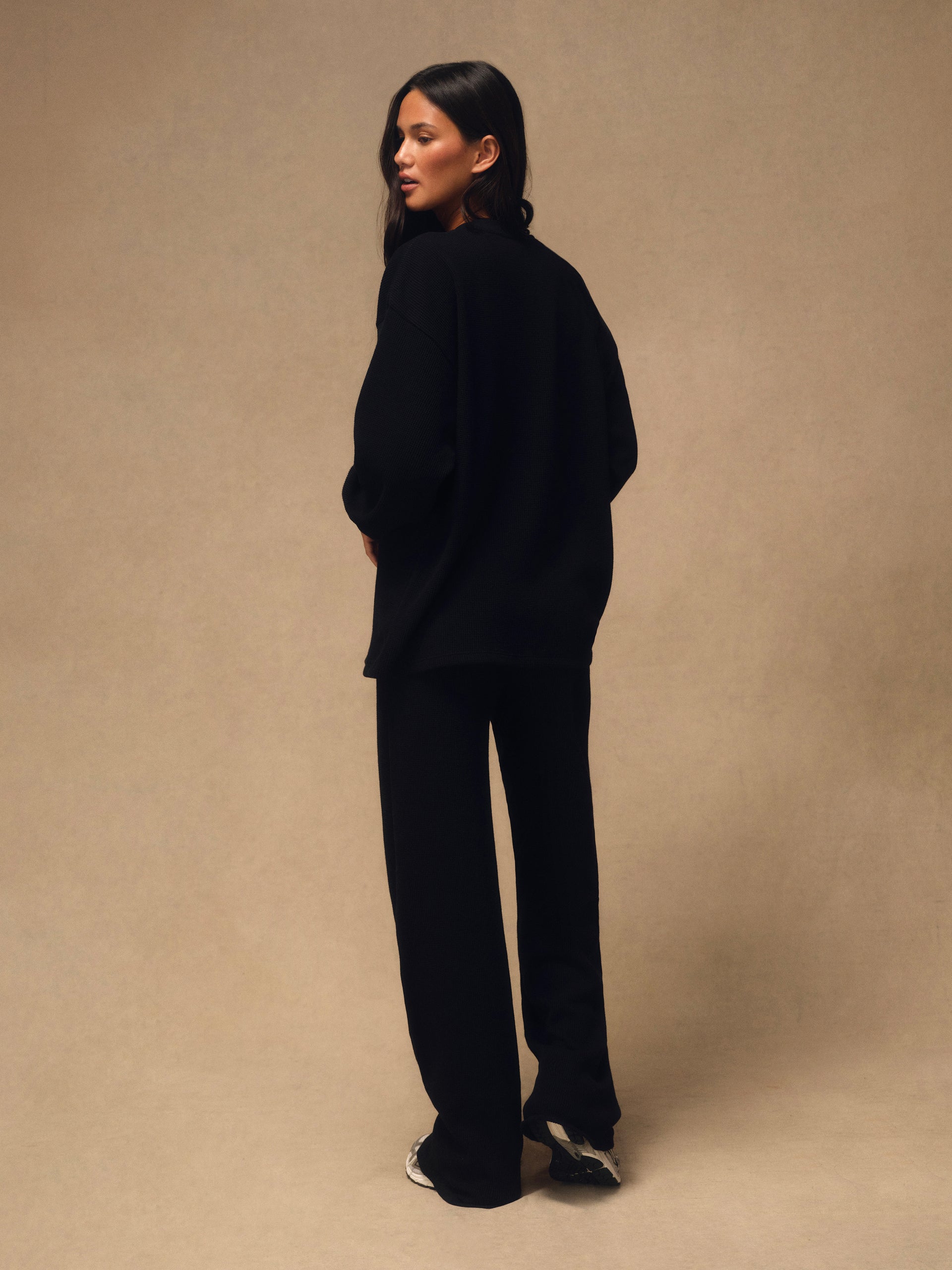 A woman stands in profile wearing an oversized black knit top and matching black wide-leg pants against a plain warm beige studio backdrop.