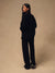 A woman stands in profile wearing an oversized black knit top and matching black wide-leg pants against a plain warm beige studio backdrop.