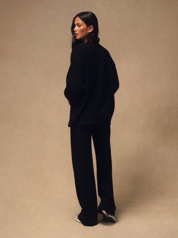 A woman stands in profile wearing an oversized black knit top and matching black wide-leg pants against a plain warm beige studio backdrop.
