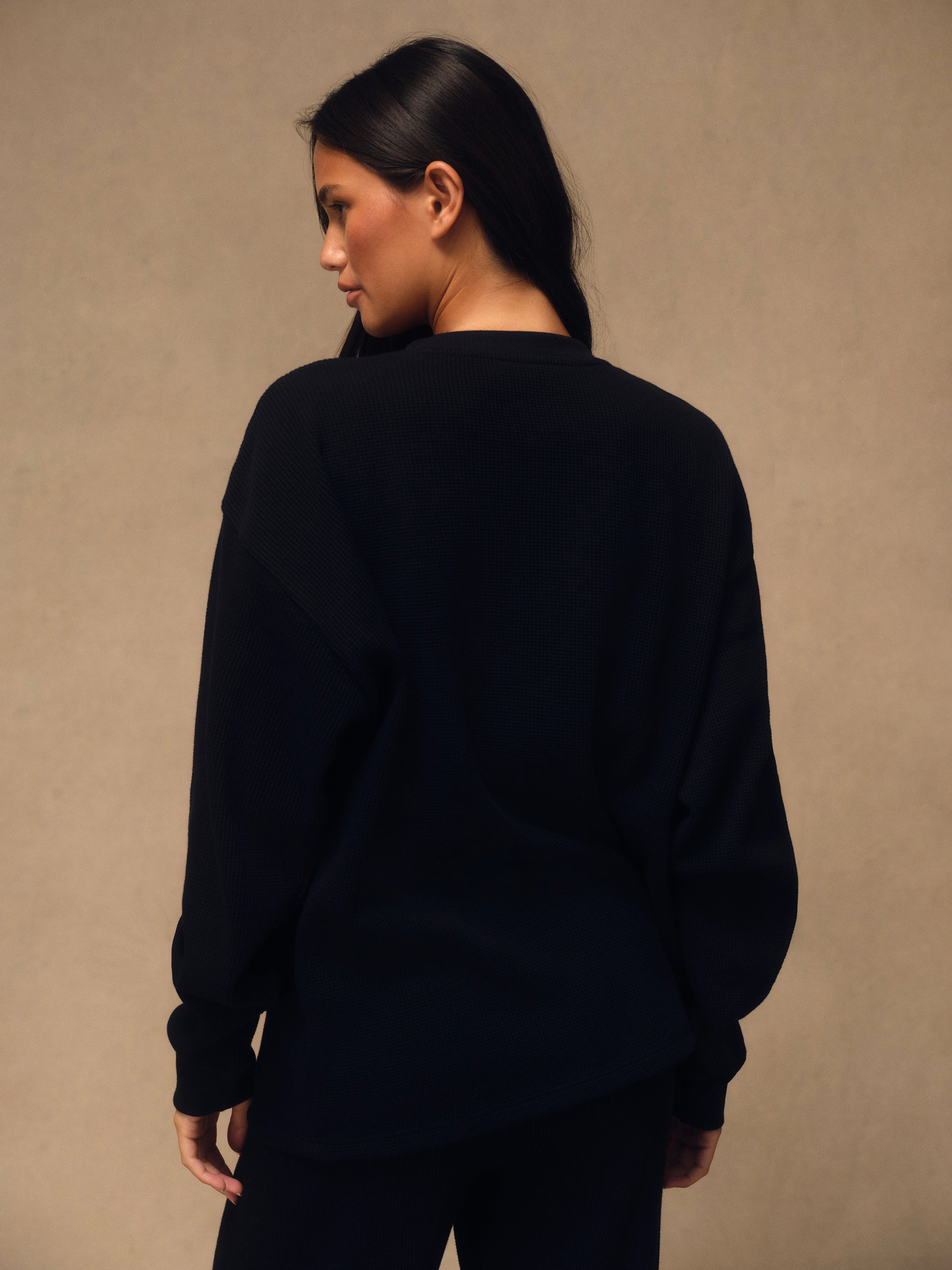 A woman with long dark hair is shown from behind wearing an oversized black crewneck sweatshirt and black pants, standing against a textured beige studio wall.