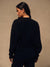 A woman with long dark hair is shown from behind wearing an oversized black crewneck sweatshirt and black pants, standing against a textured beige studio wall.