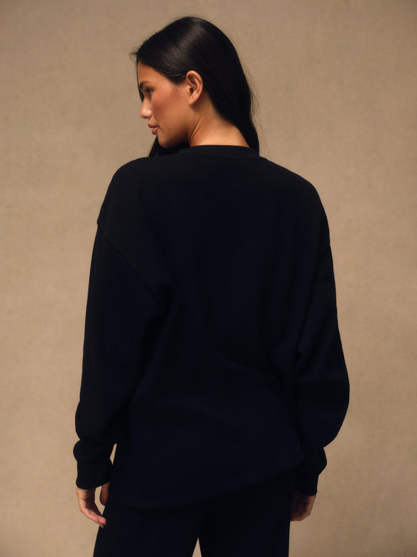 A woman with long dark hair is shown from behind wearing an oversized black crewneck sweatshirt and black pants, standing against a textured beige studio wall.