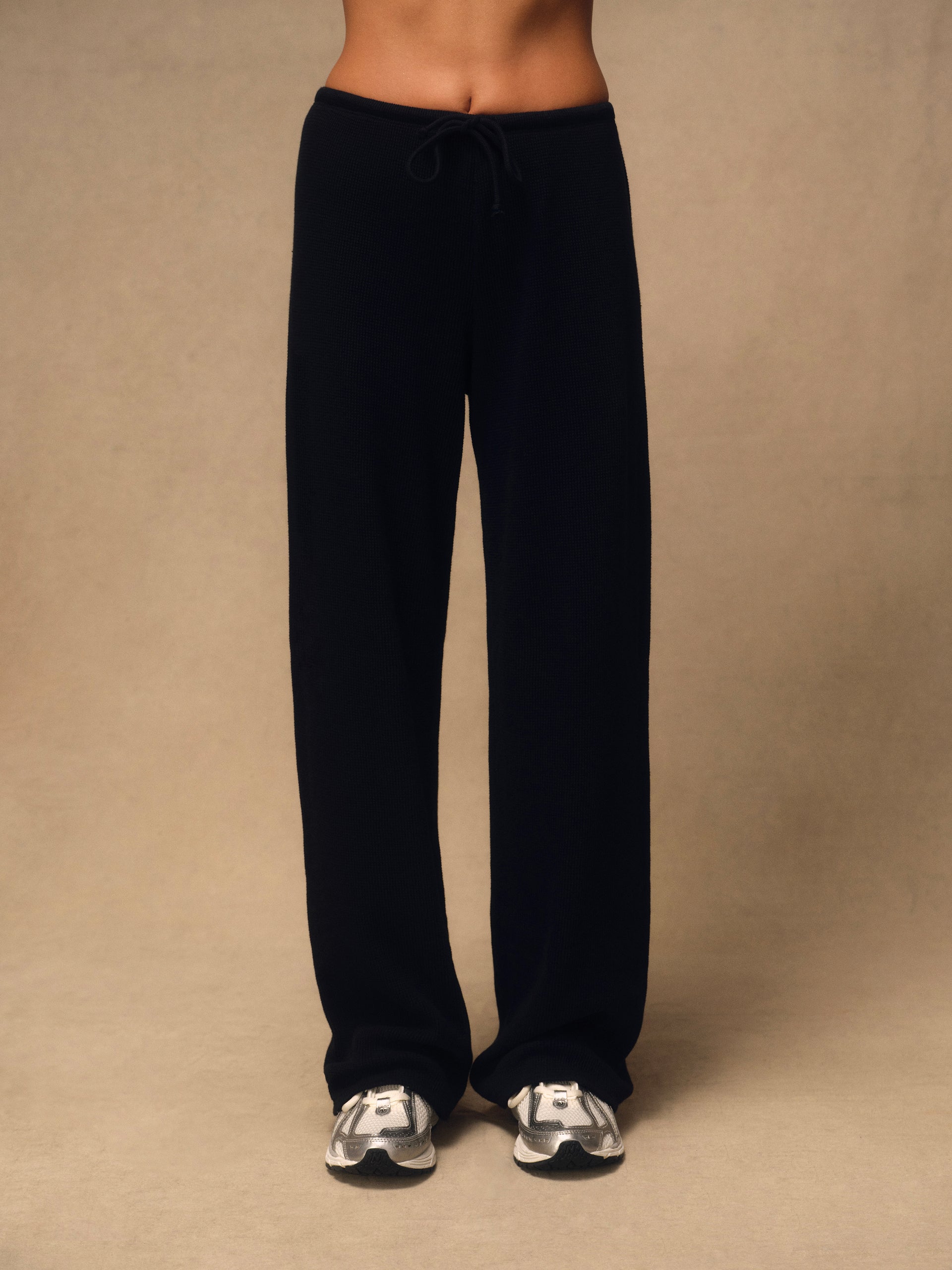 A person wearing loose black drawstring sweatpants and white sneakers, standing against a plain beige studio background.