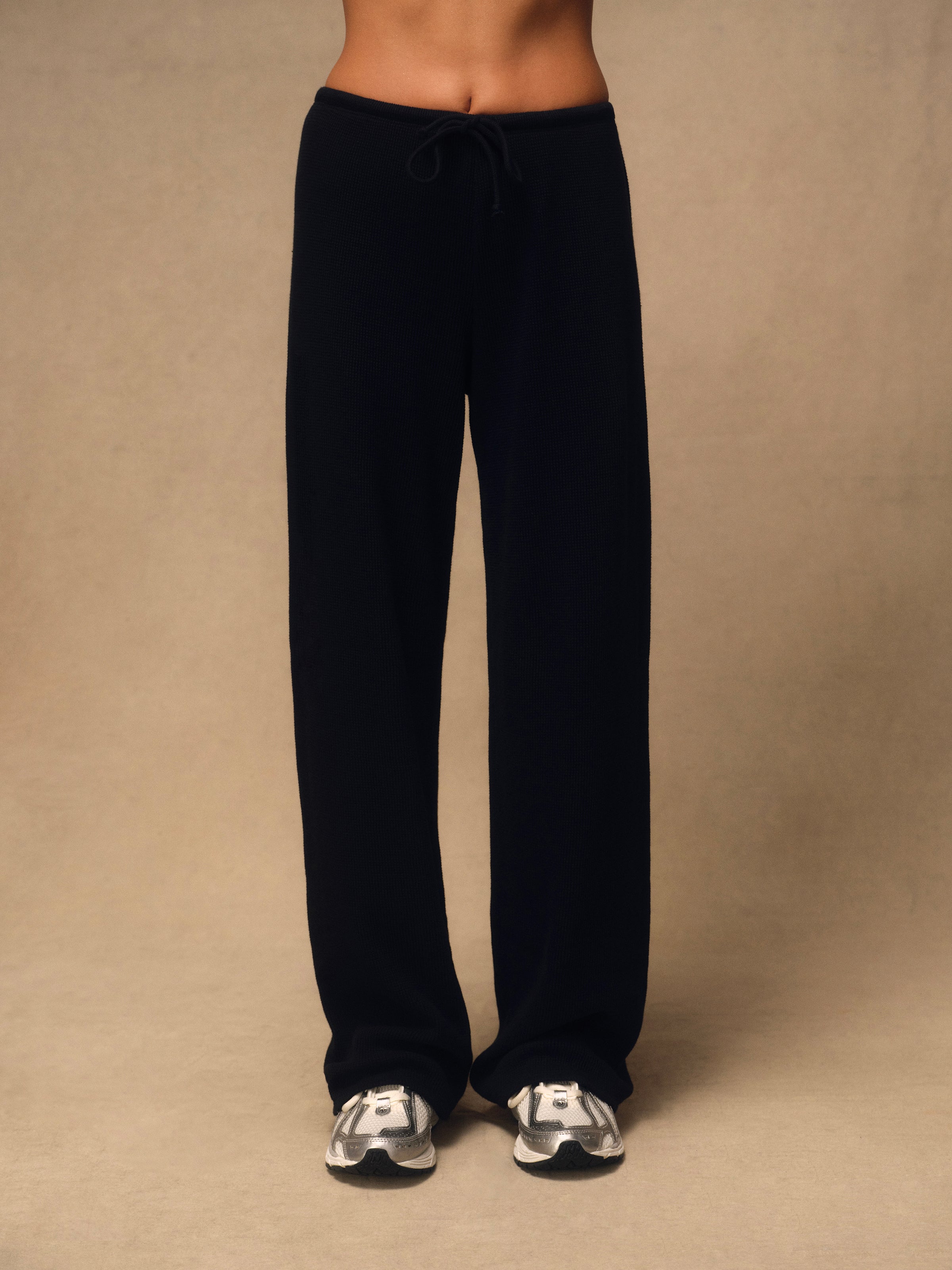 A person wearing loose black drawstring sweatpants and white sneakers, standing against a plain beige studio background.