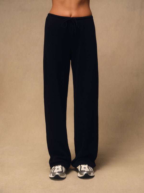 A person wearing loose black drawstring sweatpants and white sneakers, standing against a plain beige studio background.
