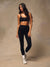 A woman modeling a matching black activewear set consisting of a sports bra and high-waisted leggings, paired with white socks and white sneakers, against a textured beige studio backdrop.
