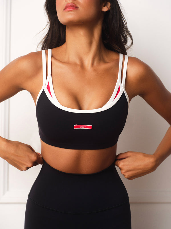 A woman wearing a black sports bra with white and red trim, paired with a matching black high-waisted sports bottom, shown against a light-colored paneled wall.