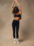 A woman wearing a black sports bra and black high-waisted leggings, standing with her arms raised overhead against a textured beige studio backdrop. <model-info>Jelene wears a size XS and is 5’7”, 23” waist, 32AB bust.</model-info>