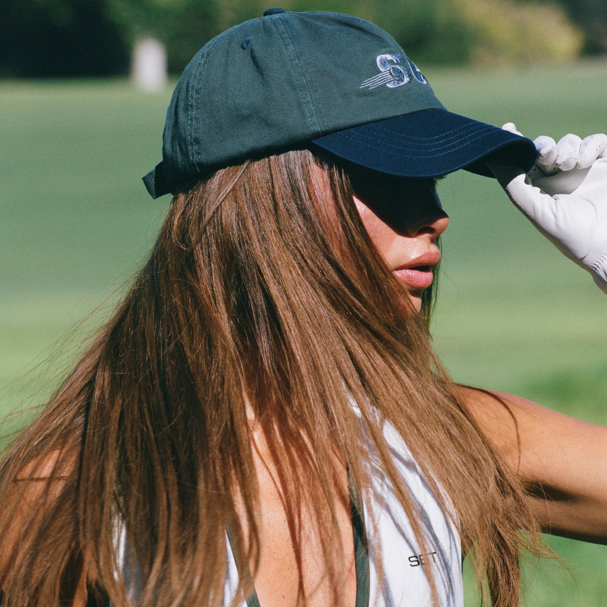 A woman on a golf course wearing a dark green baseball cap with a navy brim, white golf sunglasses, and a white glove, viewed from a side profile.