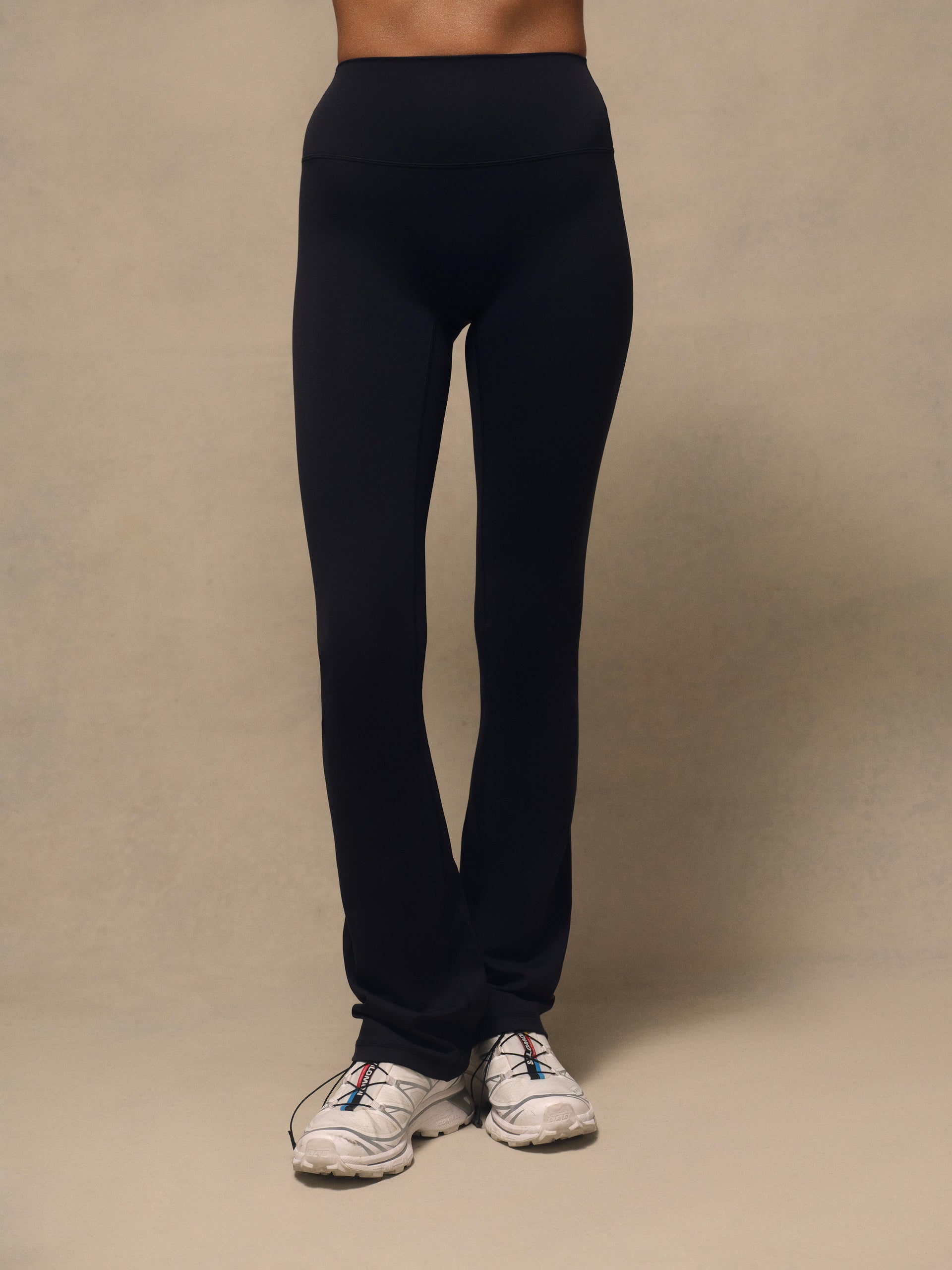 Black high-waisted athletic leggings worn by a model, paired with white sneakers, photographed against a neutral beige studio backdrop.