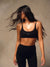 A woman wearing a black sports bra and matching black leggings, photographed against a textured beige studio background. The sports bra has a small white 'SET' logo on the front.