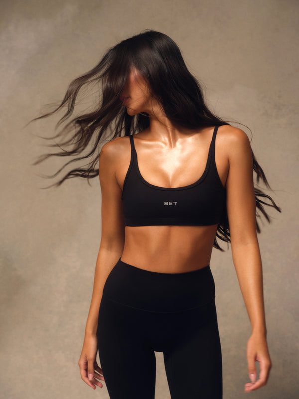 A woman wearing a black sports bra and matching black leggings, photographed against a textured beige studio background. The sports bra has a small white 'SET' logo on the front.